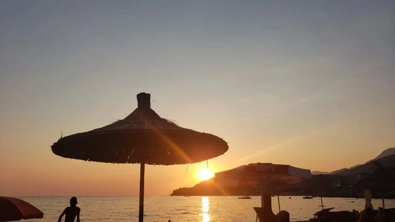 The sun dropping behind a headland, seen past the silhouette of a thatched beach umbrella