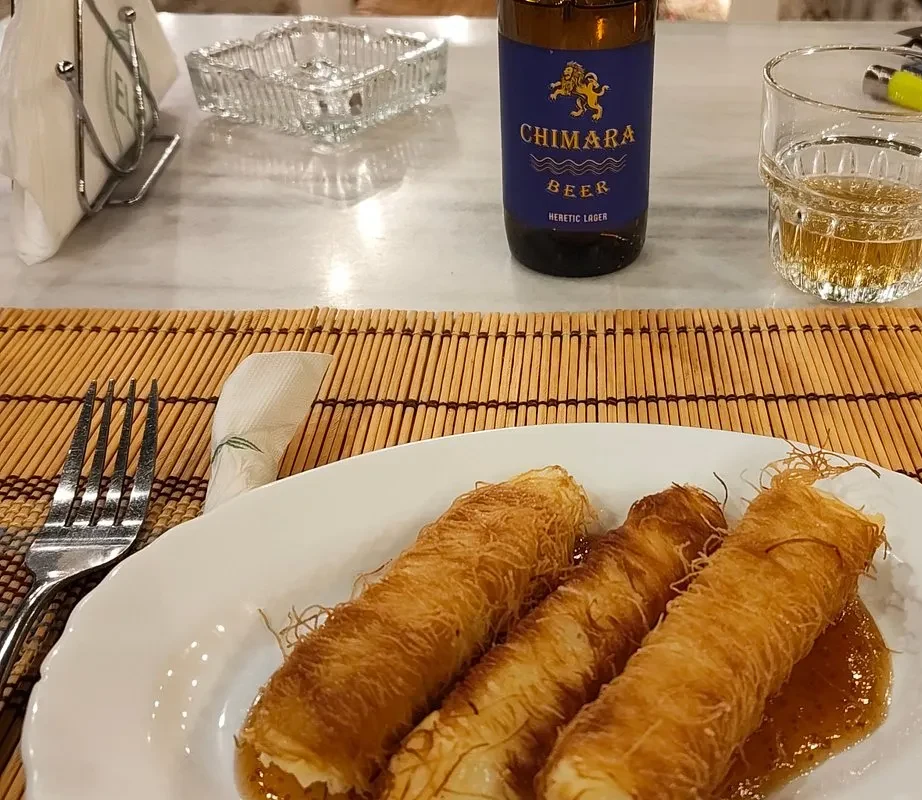 A plate of kataifi — three rolls of shredded pastry in syrup — on a marble table, beside a bottle of Chimara beer, a cut-glass tumbler of the lager, a heavy glass ashtray, a fork, a bamboo slat mat and a napkin holder carrying a pointed-oval mark