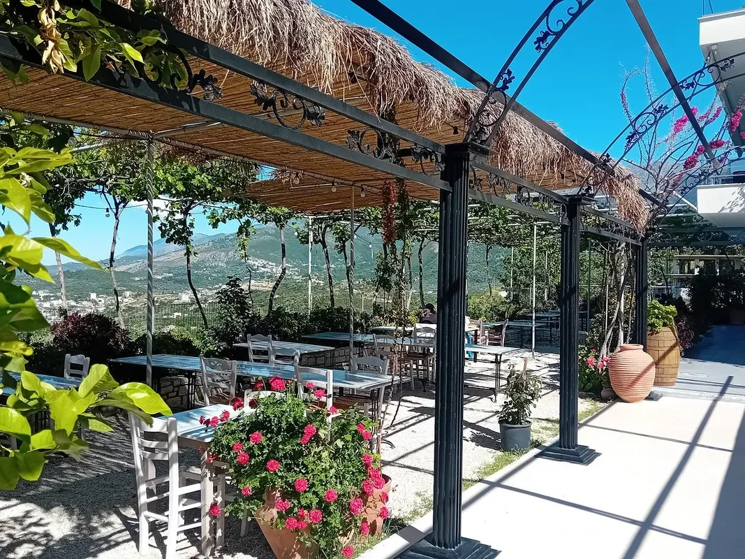 The pergola terrace: fluted black cast-iron columns carry beams hung with cast vine scrollwork, under flat cane panels, a thatch of dried grass and living grape vine, over pale blue tables and white chairs on gravel, with terracotta pots of geranium and the mountains beyond