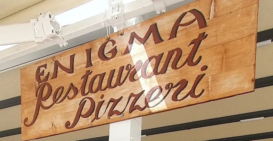 The venue's hand-painted board, hung on wires from the awning frame: ENIGMA in spaced capitals above Restaurant and Pizzeri in a brush script, dark reddish brown on bare boards