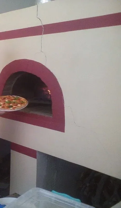The oven at Enigma: a rendered masonry dome with its arch and band painted deep red, a pizza standing on a round metal tray at the mouth, and the fire burning inside the chamber to the right of it