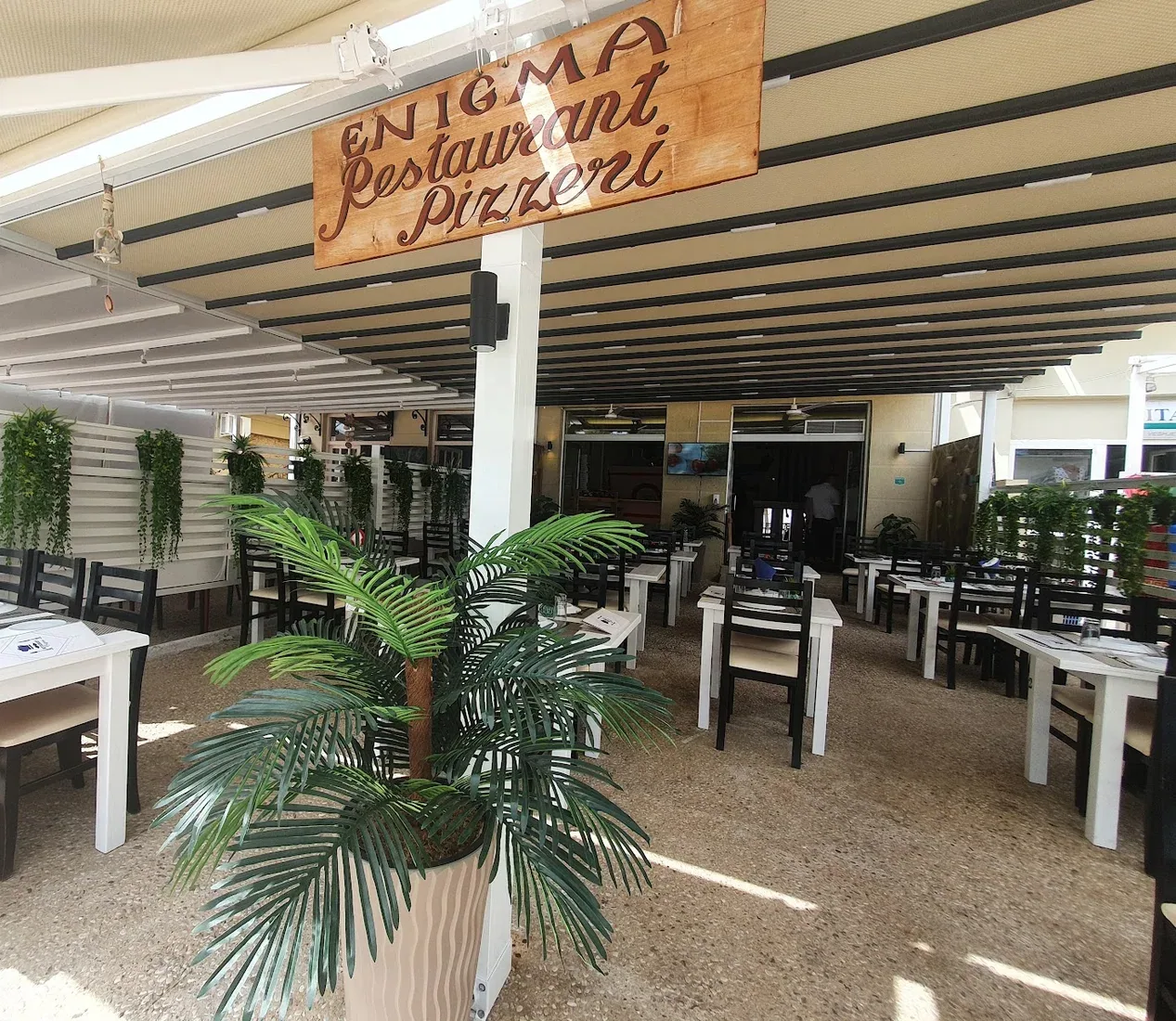 The covered terrace at Enigma: a cream retractable awning on dark battens, white tables and black slatted chairs, white slat screens hung with trailing greenery, and the painted board overhead
