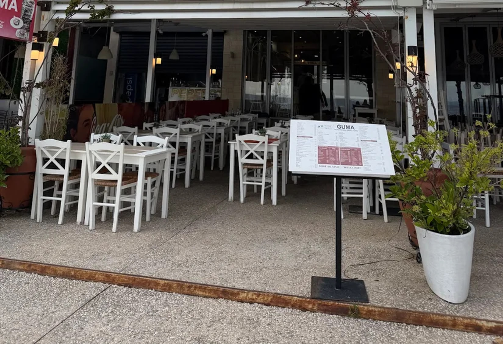 The open front seen from the promenade out of season, white cross-back chairs tipped in against cleared tables and a standing menu board out on the paving