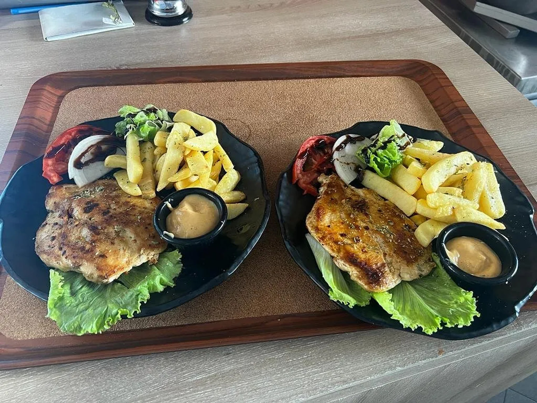 Two black plates on a cork-faced tray in a timber frame, flat seared cutlets, thick-cut fries, a tomato slice and a raw onion segment under balsamic, dressed leaves and a ramekin of tan sauce