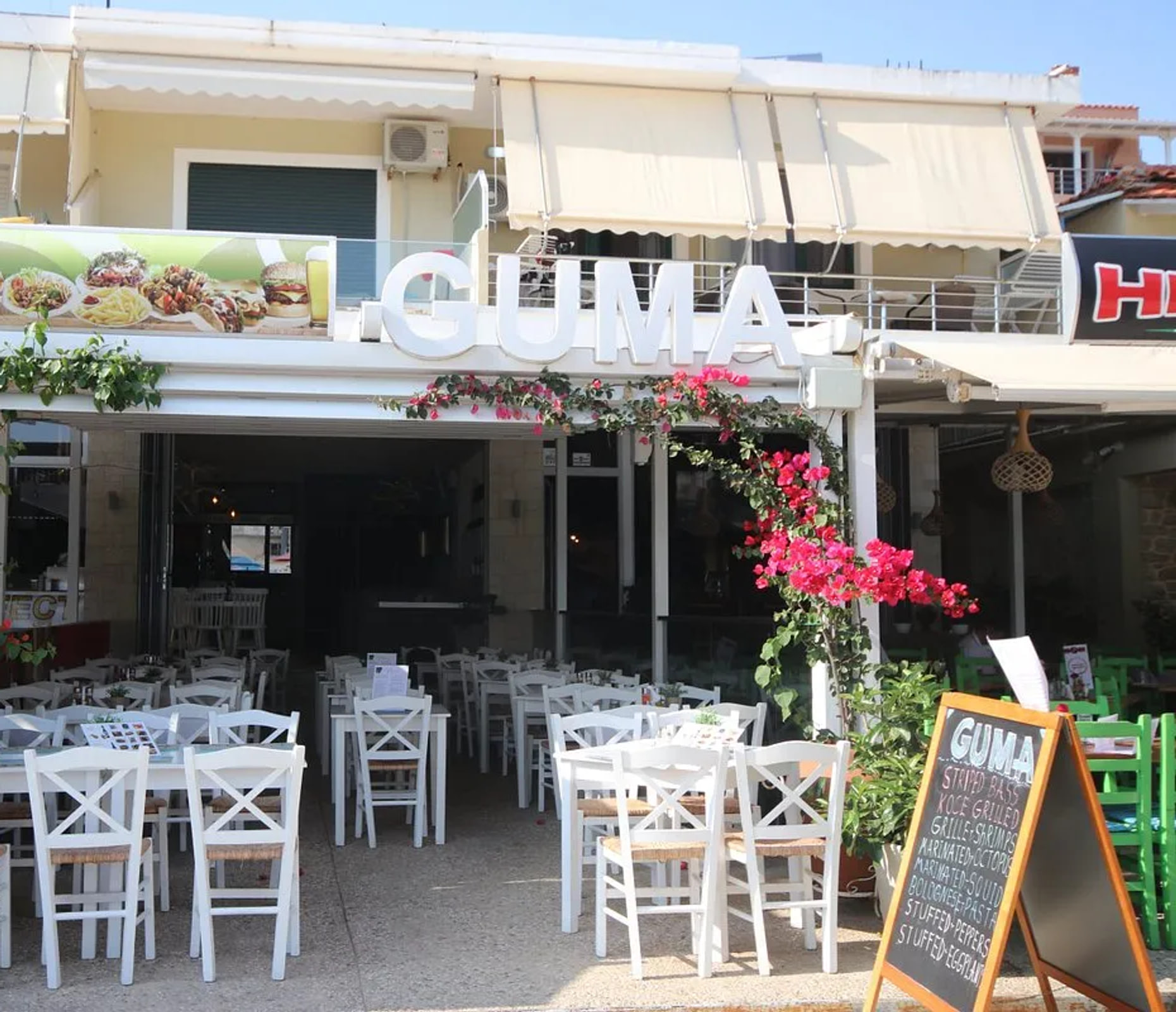 The restaurant front in summer, white channel letters reading GUMA above an open shopfront, bougainvillea trained along the pergola and white cross-back chairs set out on the pavement