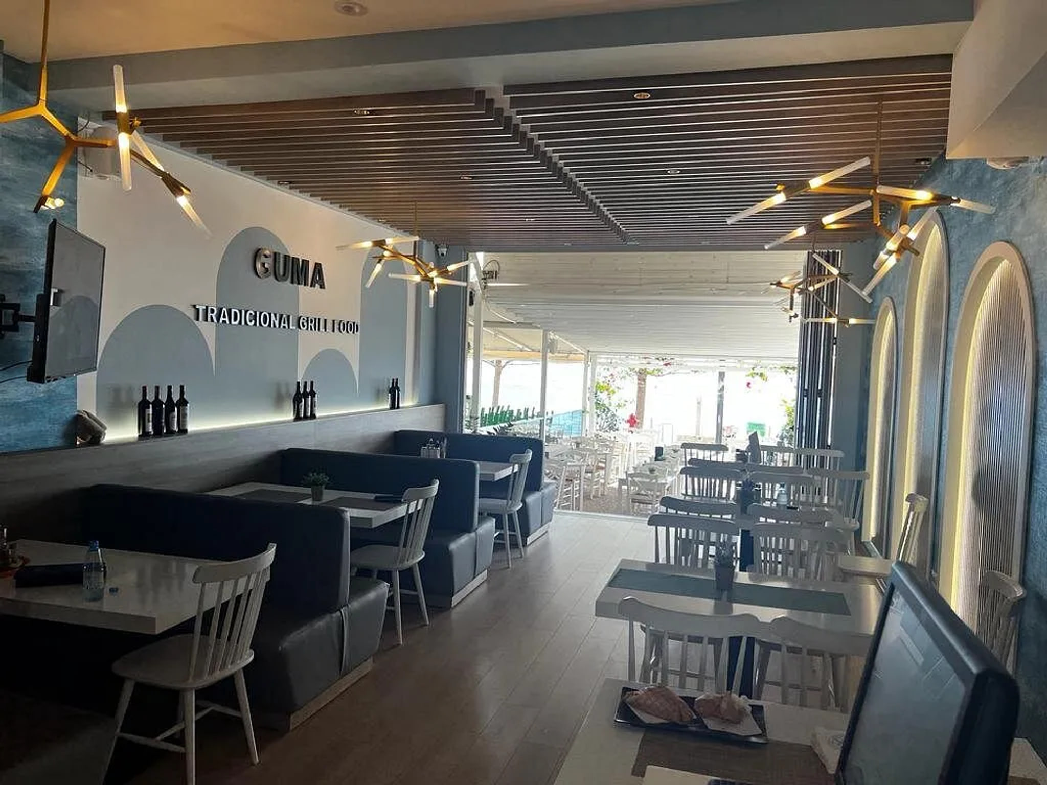 The dining room, navy banquettes and pale spindle-back chairs under a fine timber-batten ceiling and brass fittings, the front folded fully open onto the pavement terrace