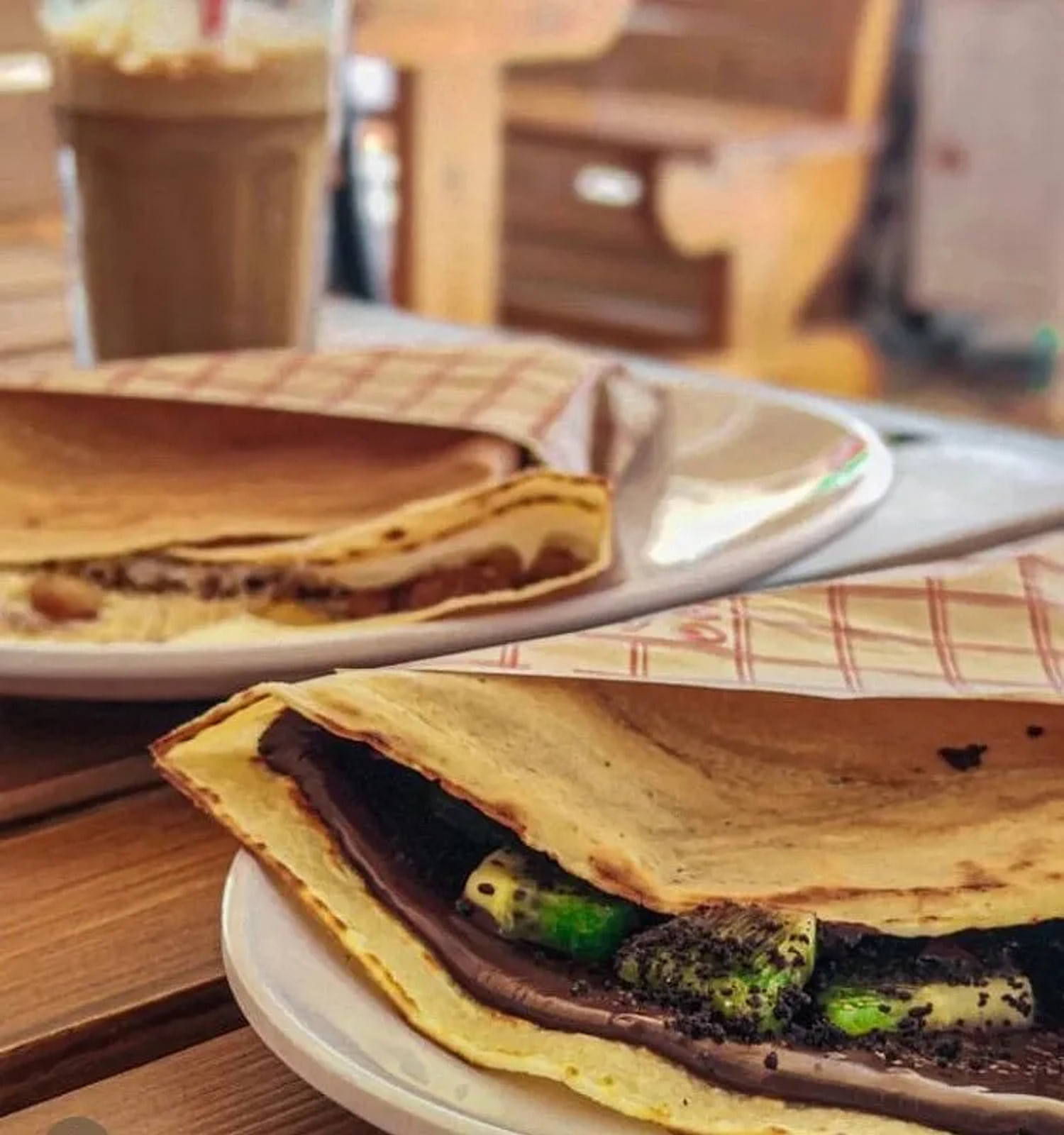 A crepe folded over chocolate spread, sliced kiwi and a scattering of dark crumb, on a white plate on a timber table, a second folded crepe on a plate behind it.