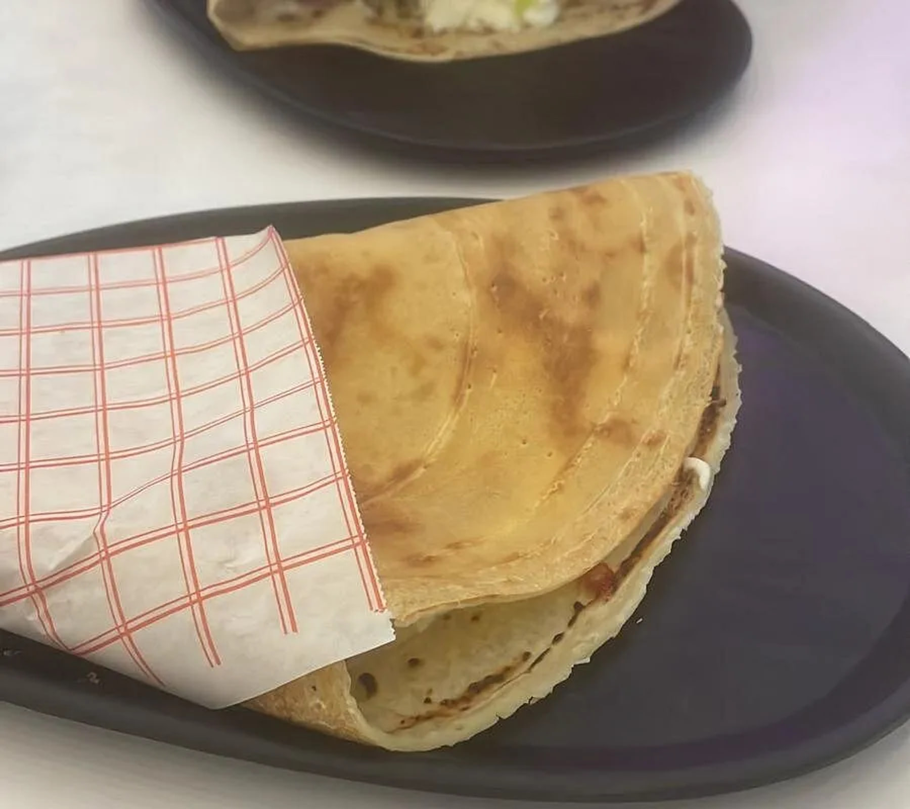 A crepe folded into quarters and wrapped halfway in greaseproof paper printed with a coral grid, on a dark plate, a second plated crepe behind it.