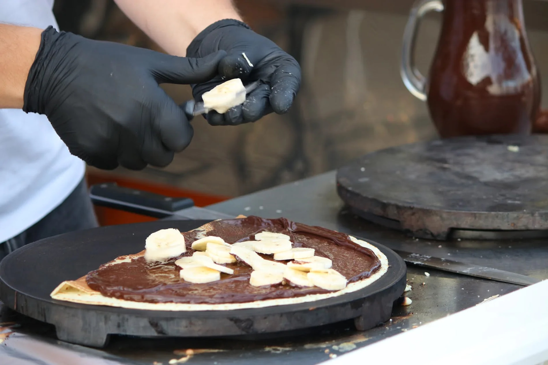 A crepe on a round black griddle, chocolate spread across it and sliced banana laid on top by two gloved hands, a glass jug of chocolate standing on a second griddle plate behind.