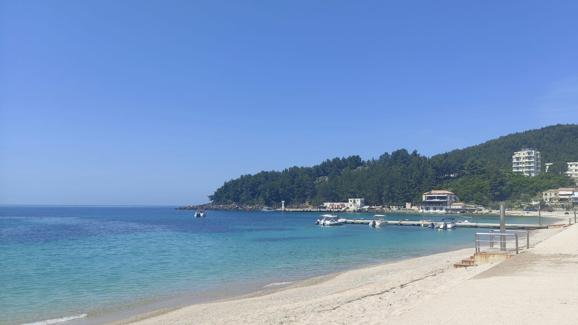 Spile beach at Himarë looking along the bay to the jetty, its moored boats and the pine-covered headland beyond