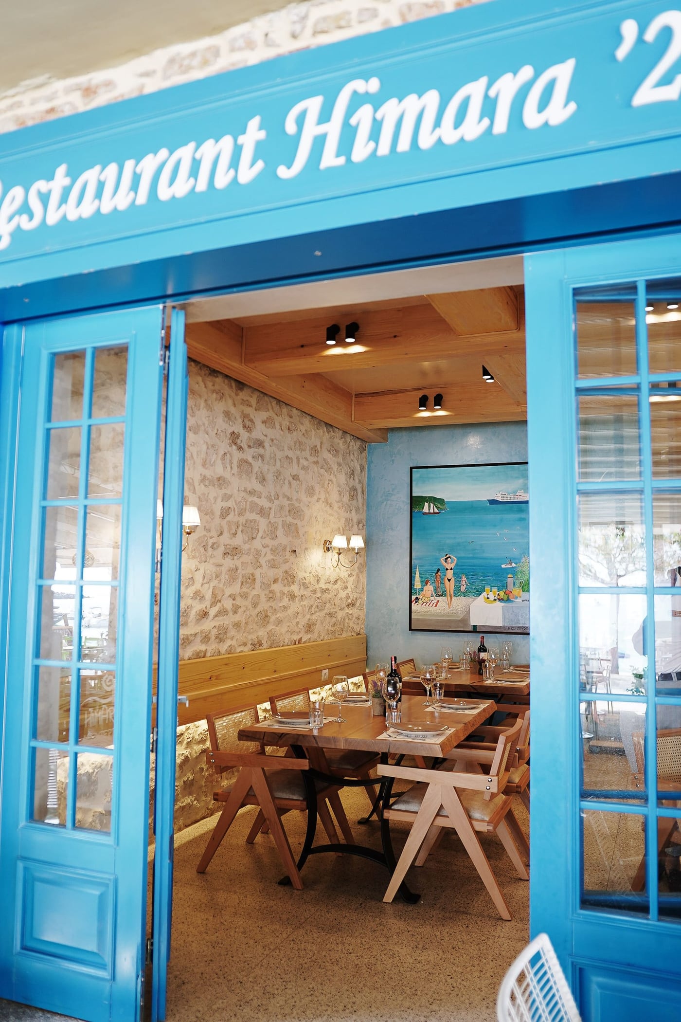 Open blue folding doors under a fascia lettered Restaurant Himara '28, looking into a dining room of stone, pale timber and cane chairs