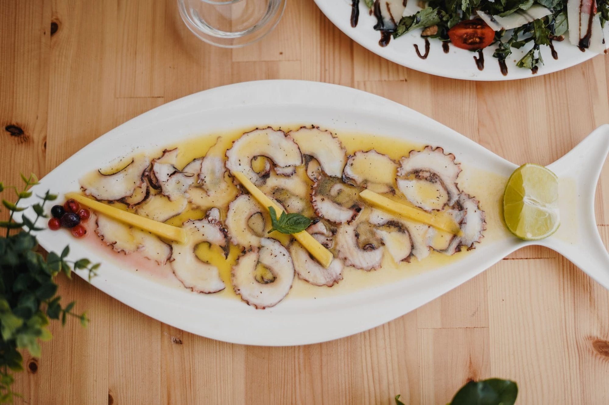 Thinly sliced octopus in oil and lemon along a white fish-shaped dish, with lime at the tail