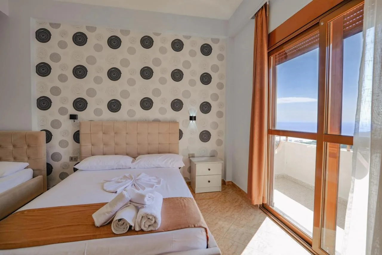 A bedroom with circle-motif wallpaper and a panel-stitched tan headboard, its wood-toned balcony door looking past the balcony rail and a strip of hillside to open sea and a clean horizon.
