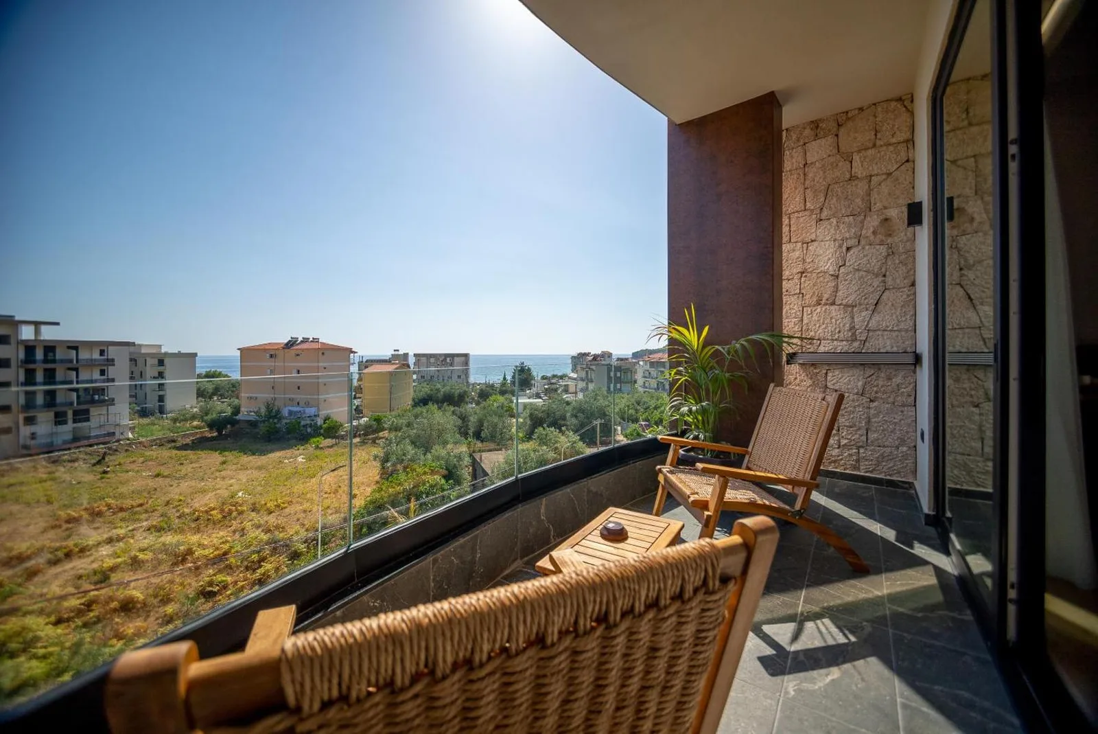 A balcony at Hotel Potami with two woven timber chairs and a low table, a stone-clad wall to one side and the Ionian showing between the apartment blocks below