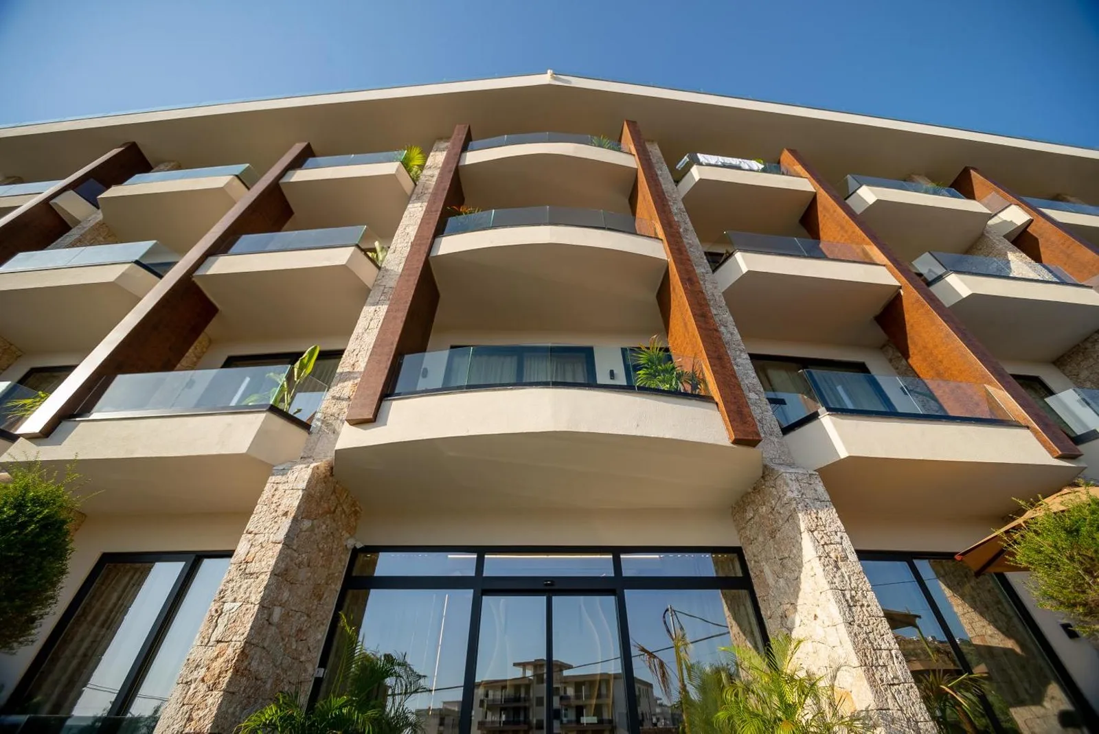 Looking up the entrance elevation of Hotel Potami: a glazed ground floor between split limestone piers, with corten steel fins running the three floors above and stopping on the first-floor slab
