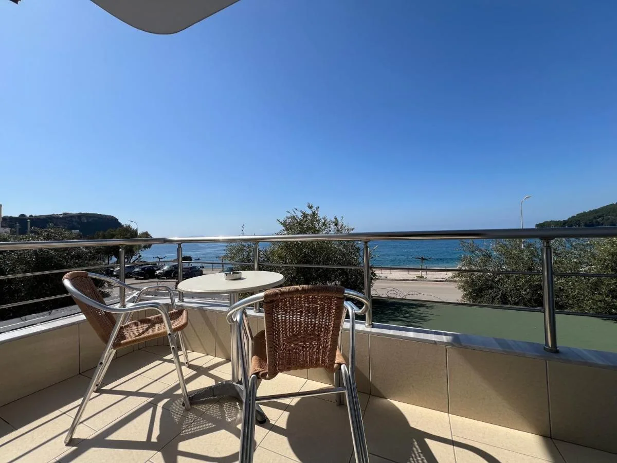 A balcony at Hotel Prinos with a round table and two wicker chairs, looking over the road and the beach to the sea, with a headland on the left horizon