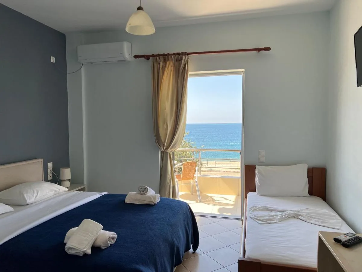 A room at Hotel Prinos with a double bed under a navy blanket, a single bed beside it, and an open balcony door showing the sea