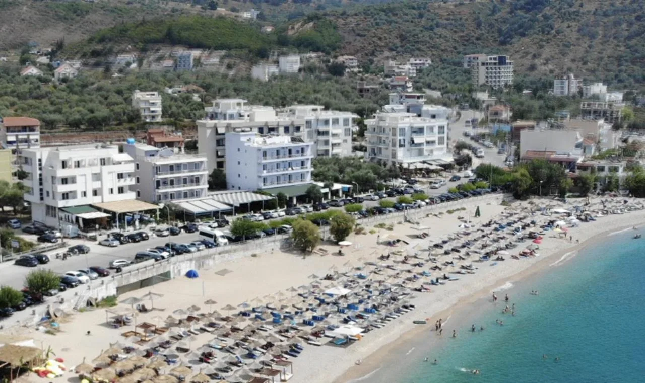 The Potam strip photographed from the air: a row of white blocks, a line of parked cars along the road, a low sea wall, then rows of straw umbrellas on the pale beach and turquoise water