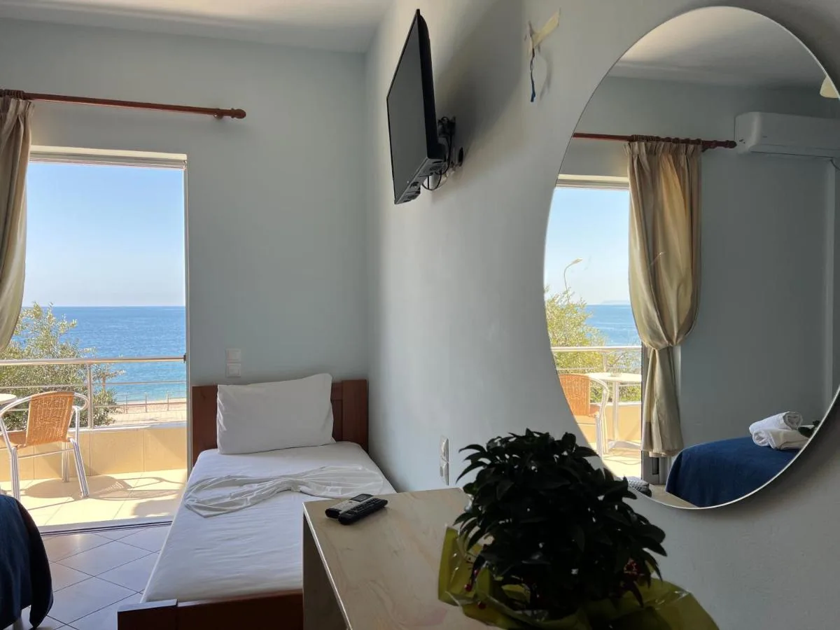 A room at Hotel Prinos where a large round mirror on the wall reflects the balcony, its rail and the sea, while the open balcony door shows the same view directly