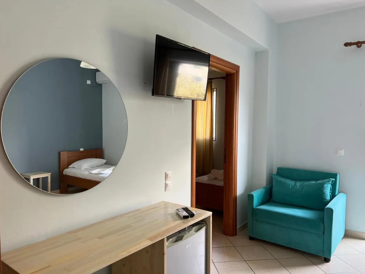 A round mirror above a pale timber counter at Hotel Prinos, reflecting a made bed in the adjoining room, with a teal armchair and a doorway in orange-stained wood