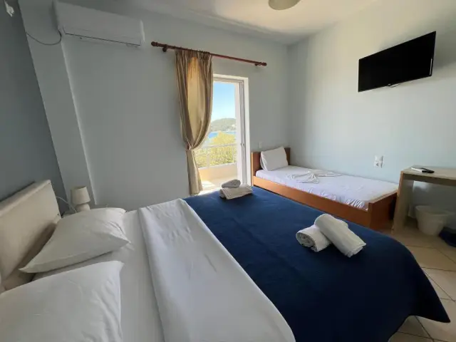 A room at Hotel Prinos with a slate-grey wall behind the beds, a double bed with a navy blanket, a single bed with a wooden frame, and a balcony door with the sea beyond