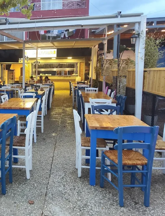 Blue-painted tables with bare wood tops and white and blue rush-seat chairs under the white pergola at I Love Souvlaki, the lit grill counter at the back