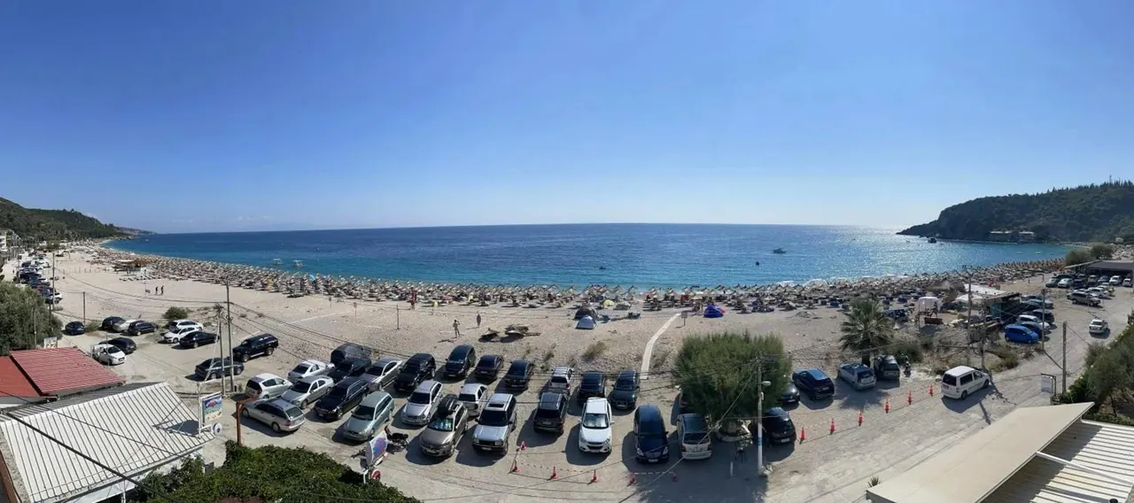 Livadhi Beach photographed from the property: the beach road and the car park in front, then rows of sun umbrellas and the bay curving to the headland