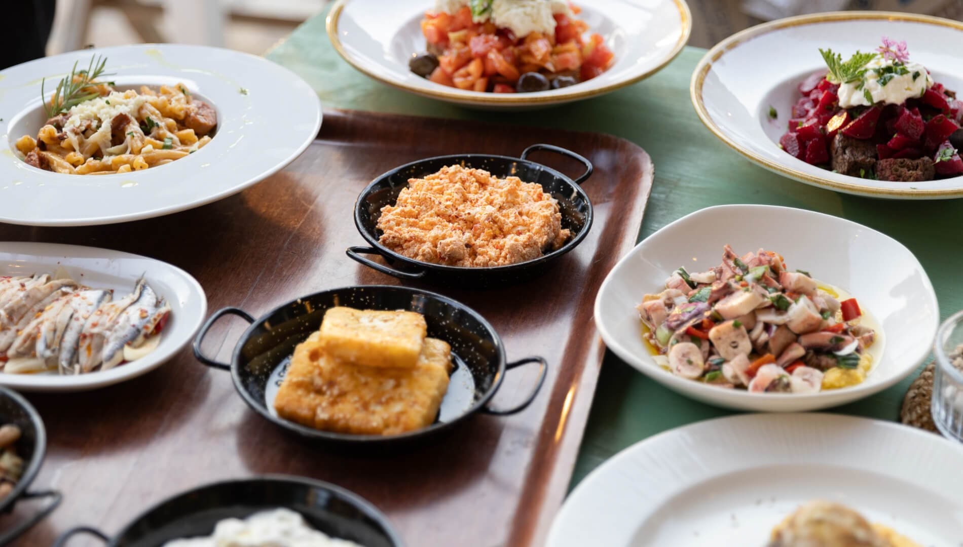A painted table laid with plates of pasta, tomato salad, beetroot and marinated anchovies, black enamel pans of fried cheese and of a coral-coloured dip, and a bowl of chopped octopus