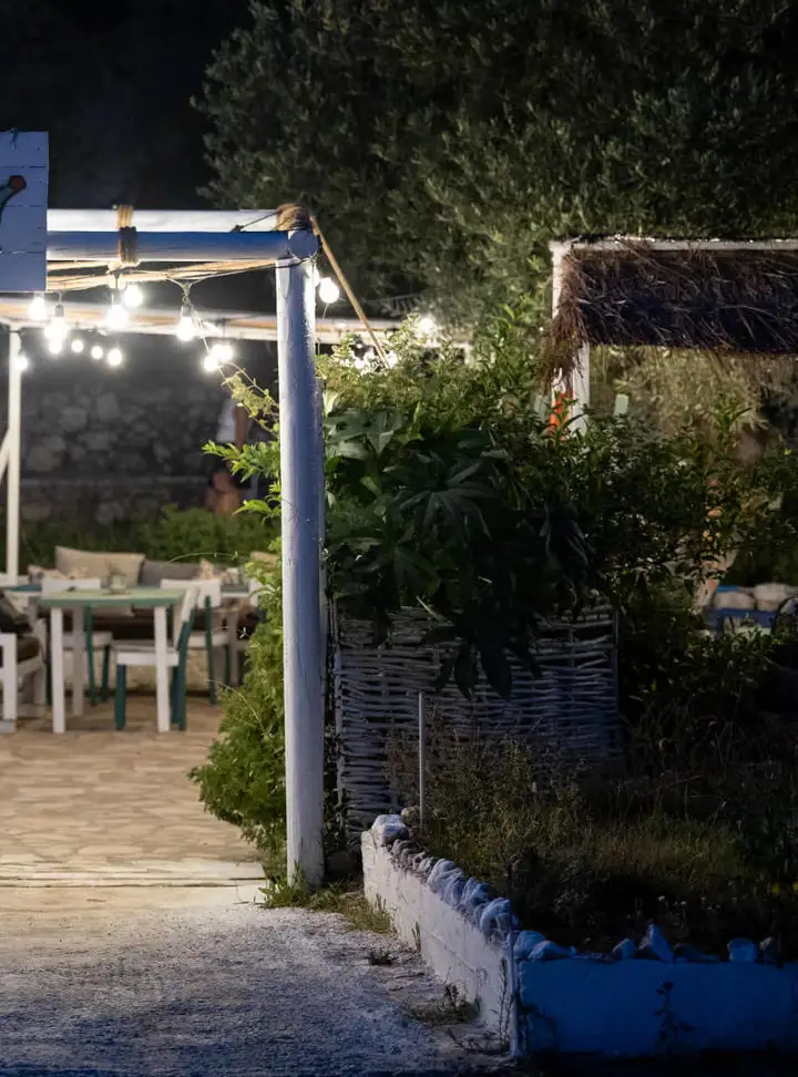 The taverna garden at night: festoon bulbs strung under a white timber pergola, an olive tree behind it, a white table with green-framed chairs to the left and a woven willow screen to the right