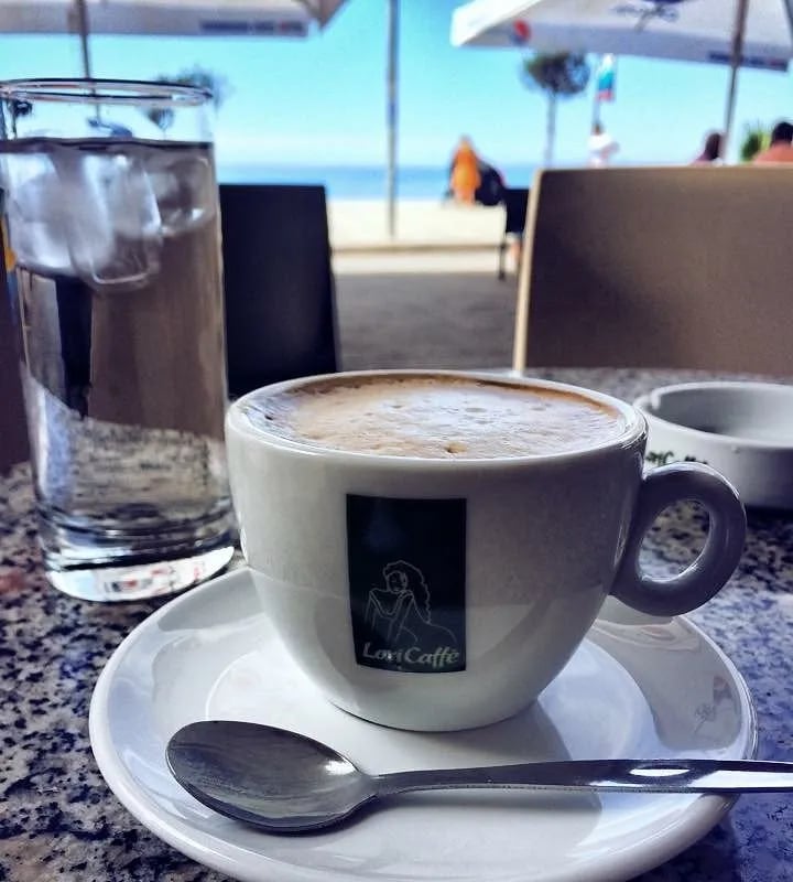 A cappuccino and a glass of iced water on a granite table at Kafe Pastiçeri Himara 1928, umbrellas up and the sea a few metres beyond