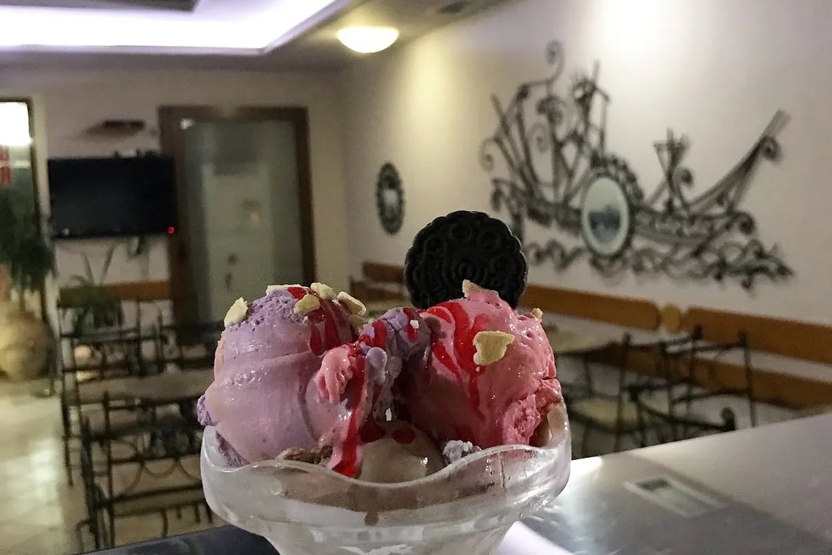 An ice cream in a glass coupe in the room at Kafe Pastiçeri Himara 1928, wrought-iron chairs behind it and a wrought-iron sailing ship fixed to the plaster wall