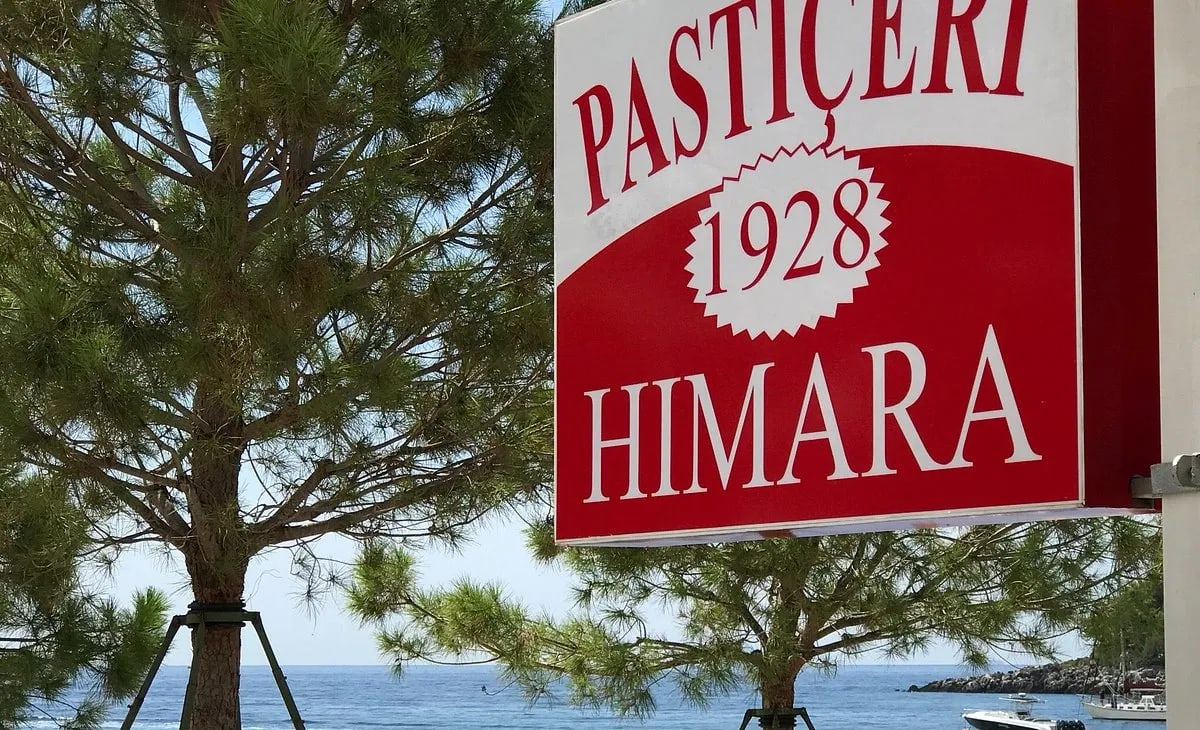 The painted board of Kafe Pastiçeri Himara 1928 on its post above the water at Spile, pines in front of it and boats moored beyond