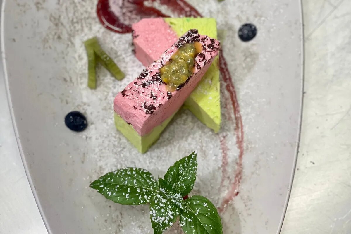A layered pink and pistachio-green dessert, dusted with sugar and topped with a spoonful of translucent yellow fruit, a sprig of basil on the plate beside it.