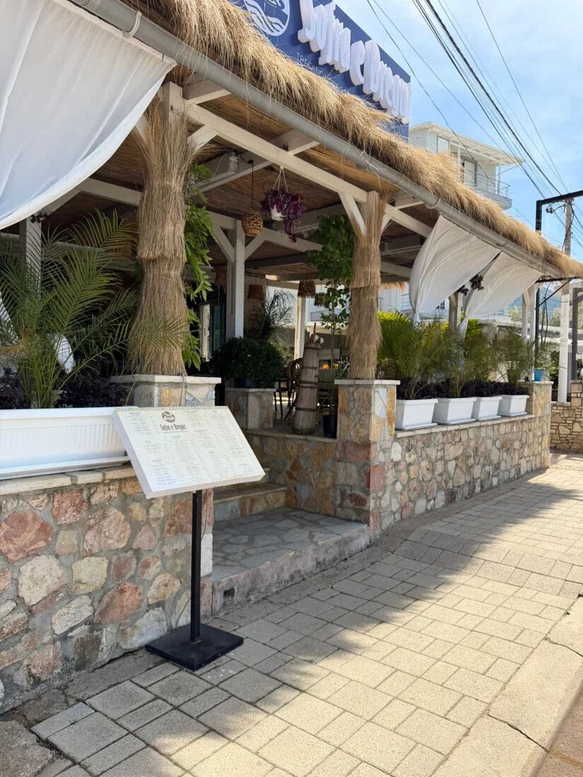 The restaurant's frontage on Rruga Vangjel Zoto: a straw-fringed pergola over a white-painted timber frame, white drapes tied back between the posts, and a stone-clad low wall with a menu board on the pavement.