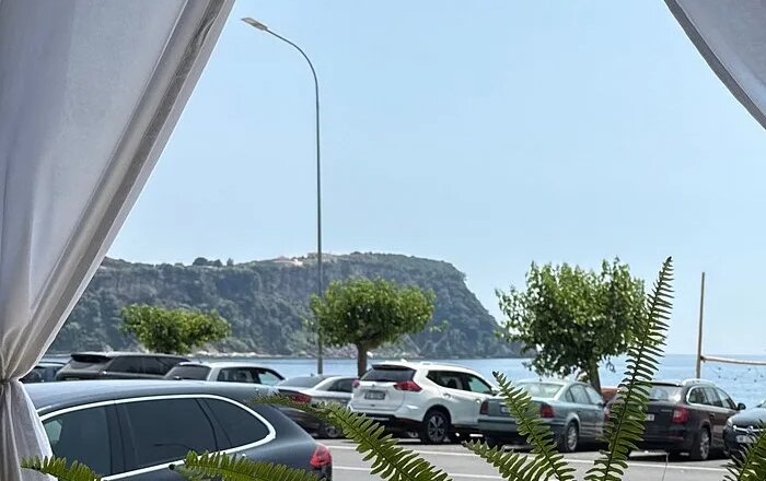 The view out from between the drapes: cars parked along the coast road, the water beyond them, and a headland standing across it.