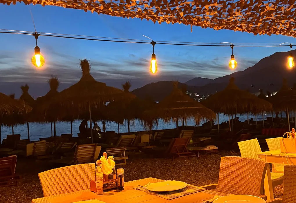Festoon bulbs under a shade net at blue hour, straw parasols and loungers on the shore, the mountains and scattered lights of the far shore behind
