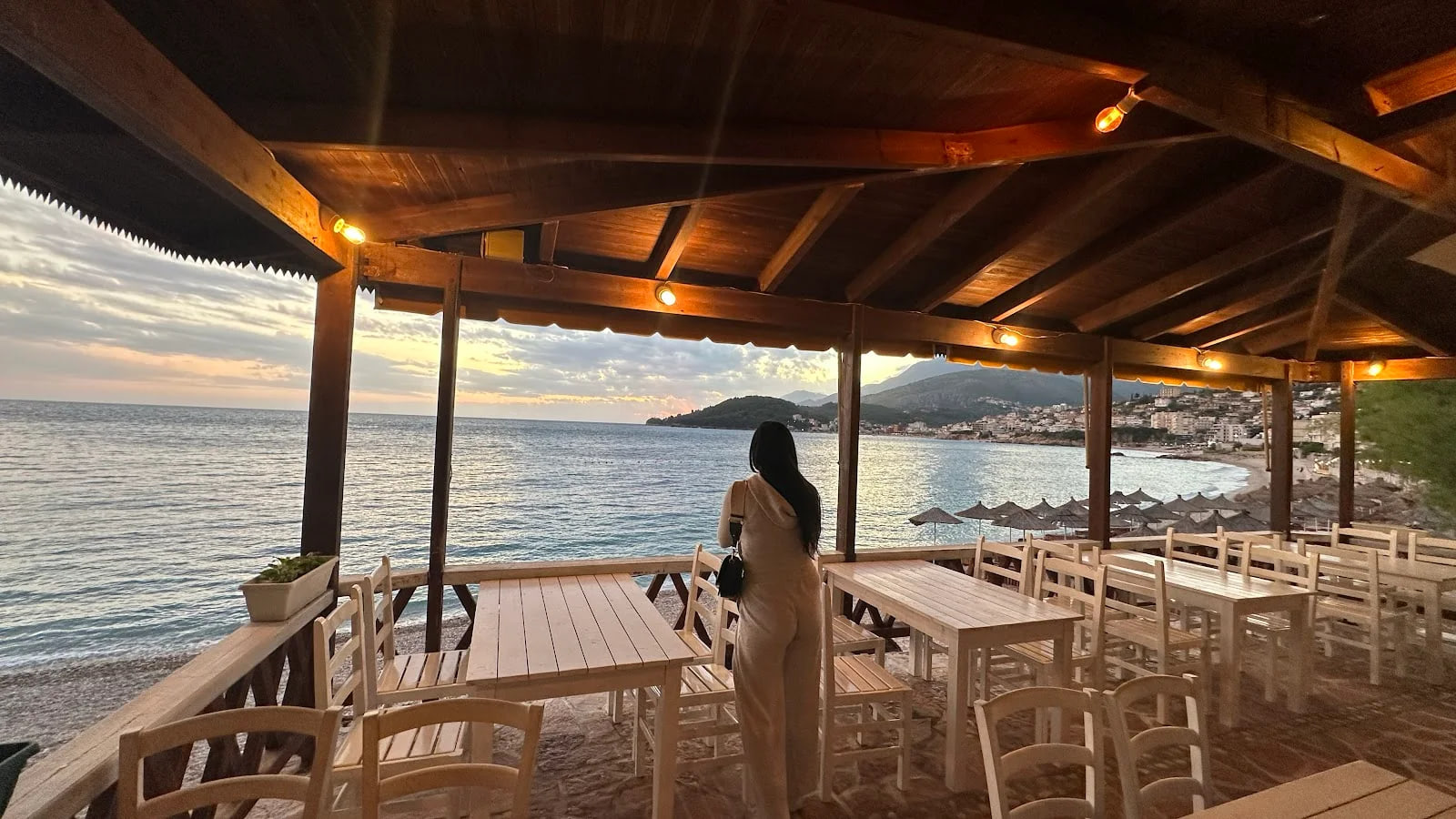 A woman stands with her back to the camera under a timber pergola, looking across the bay to the lit hillside of Himarë; pale plank tables and chairs stand on the sand around her at dusk