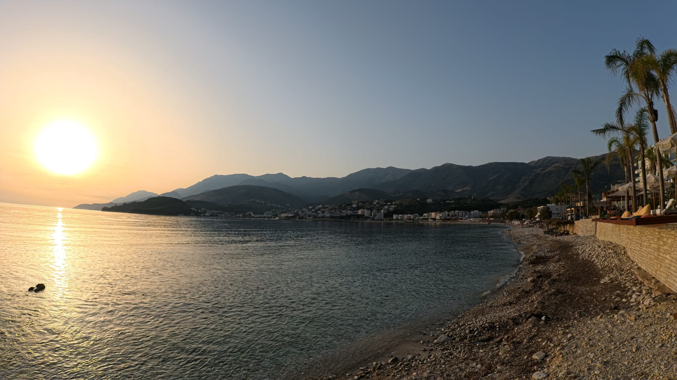 Potami Beach at sunset, the pebble shore curving away and the white buildings of Himarë along the far side of the bay