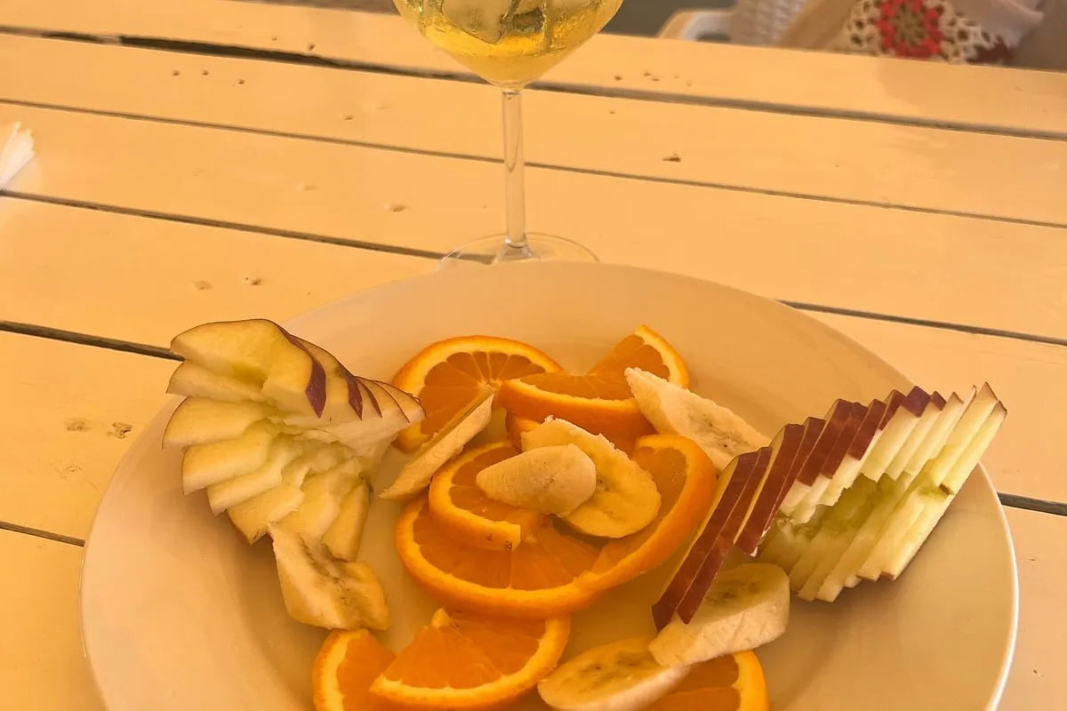 A plate of fanned apple, orange and banana beside a tall stemmed glass of an iced drink with a sprig of mint in it, on a pale timber table