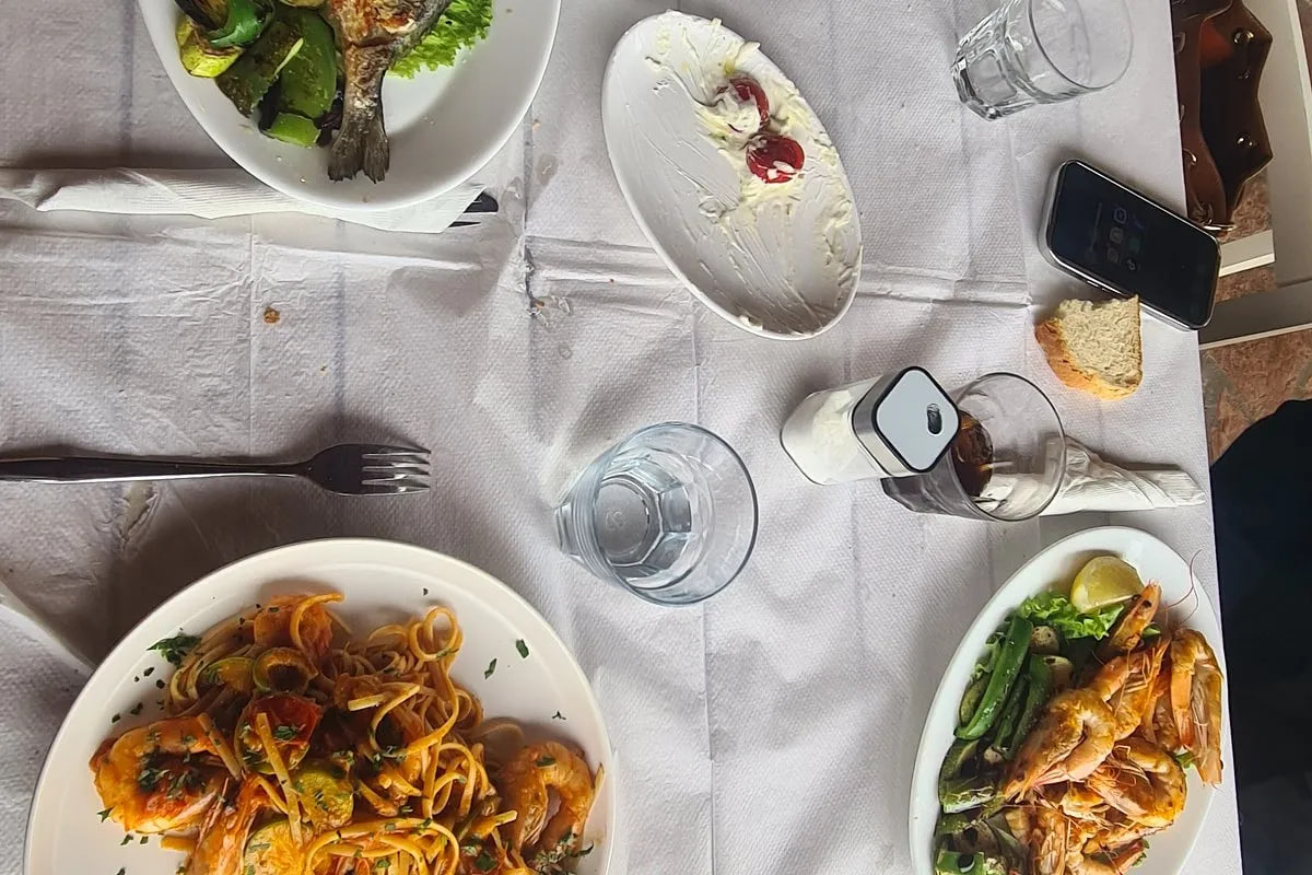 A table mid-meal seen from directly above: a grilled fish over grilled vegetables, linguine with prawns in tomato sauce, a plate of grilled prawns with lemon and a part-eaten plate of whipped cheese, on paper over the cloth