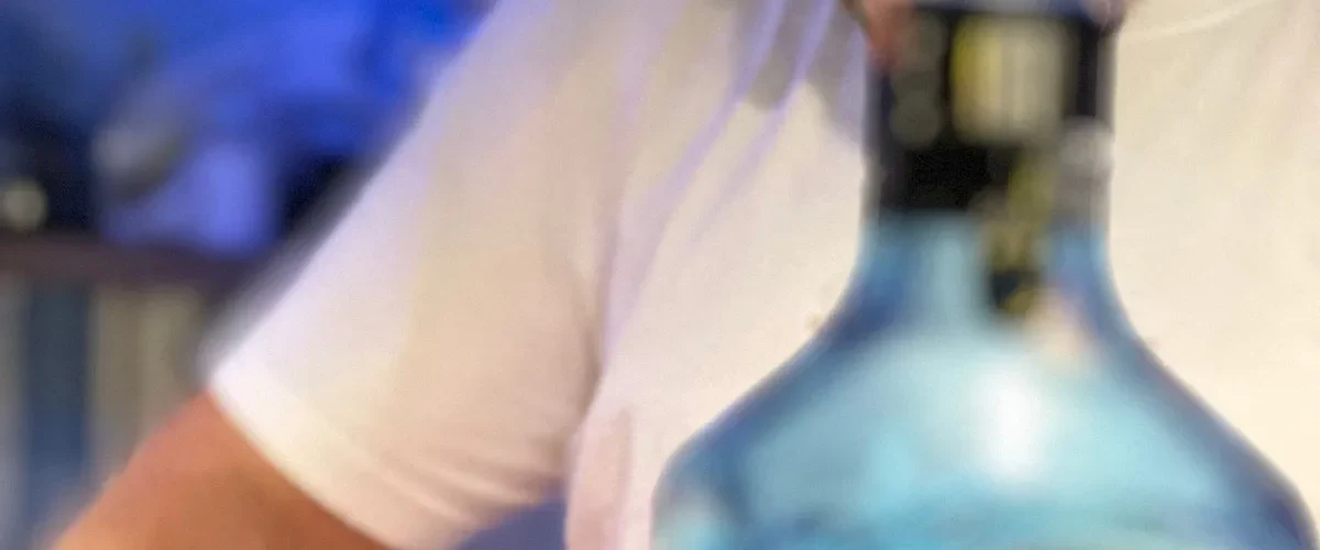 A bartender in a white T-shirt at the counter, a pale blue glass bottle standing in front of him, the room behind out of focus