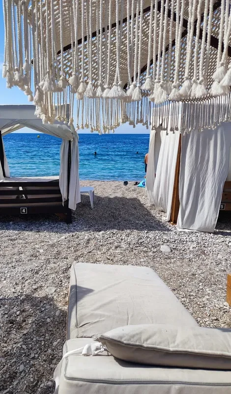 A white-curtained four-poster daybed at the top of the pebbles, with the open sea beyond