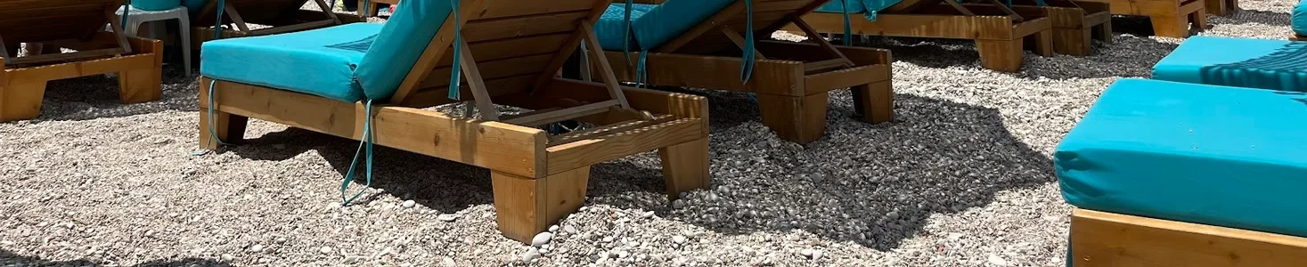 Turquoise-cushioned loungers on raw timber frames standing on the cove's white pebbles