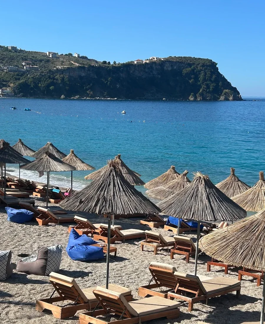 The bay at Namos Beach, Himarë: reed umbrellas over wooden loungers on grey pebbles, turquoise water, and a wooded headland closing the far side