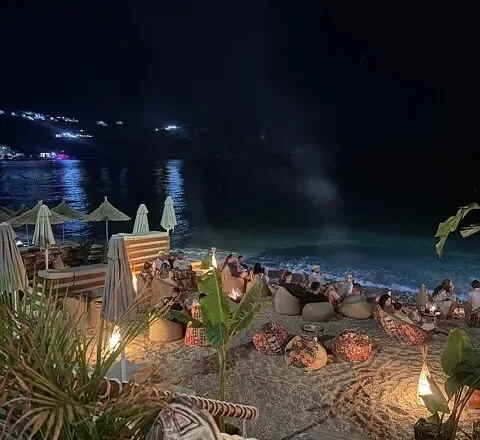Namos Beach after dark: low-lit seating on the pebbles and the lights of the far shore across the water