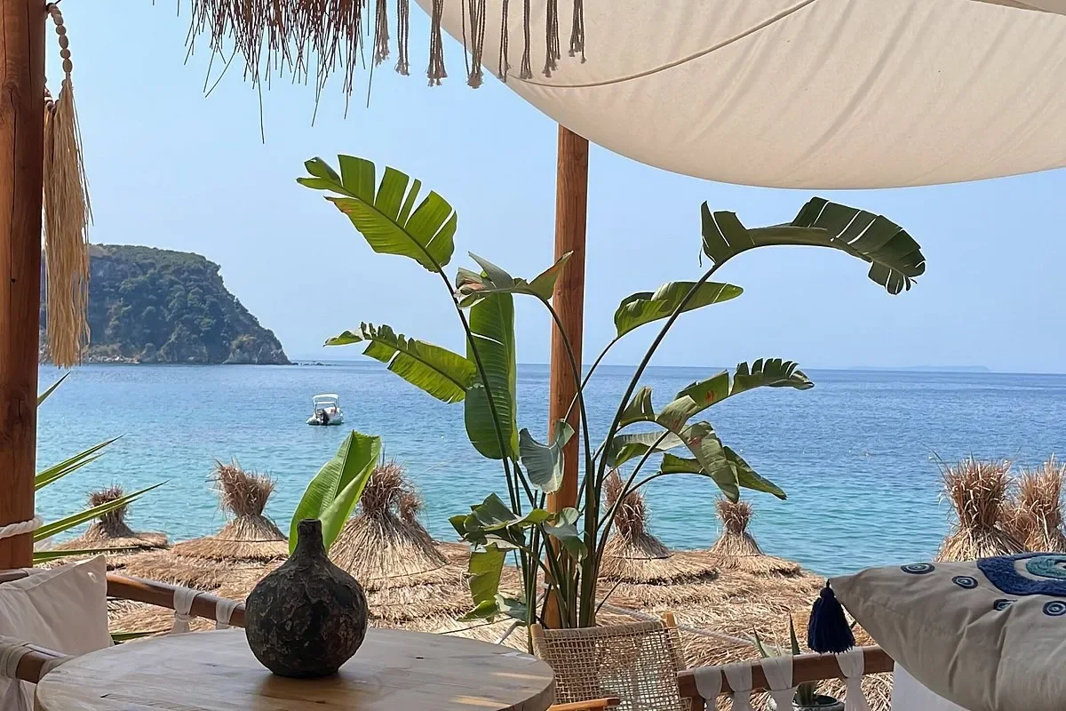 A low table under a stretched canvas sail at Namos Beach, with cushions, a banana plant and the umbrella line and sea beyond
