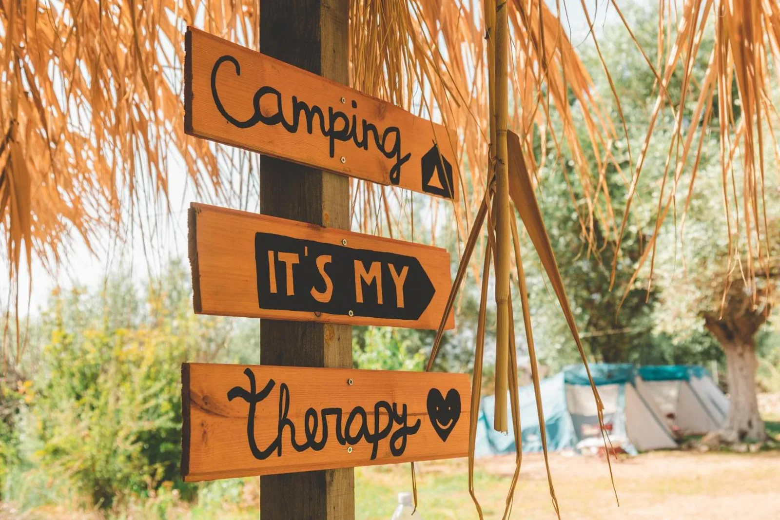 A hand-painted wooden finger-post at Olive Camping reading Camping, It's My, Therapy, under a fringe of dried palm thatch with tents behind