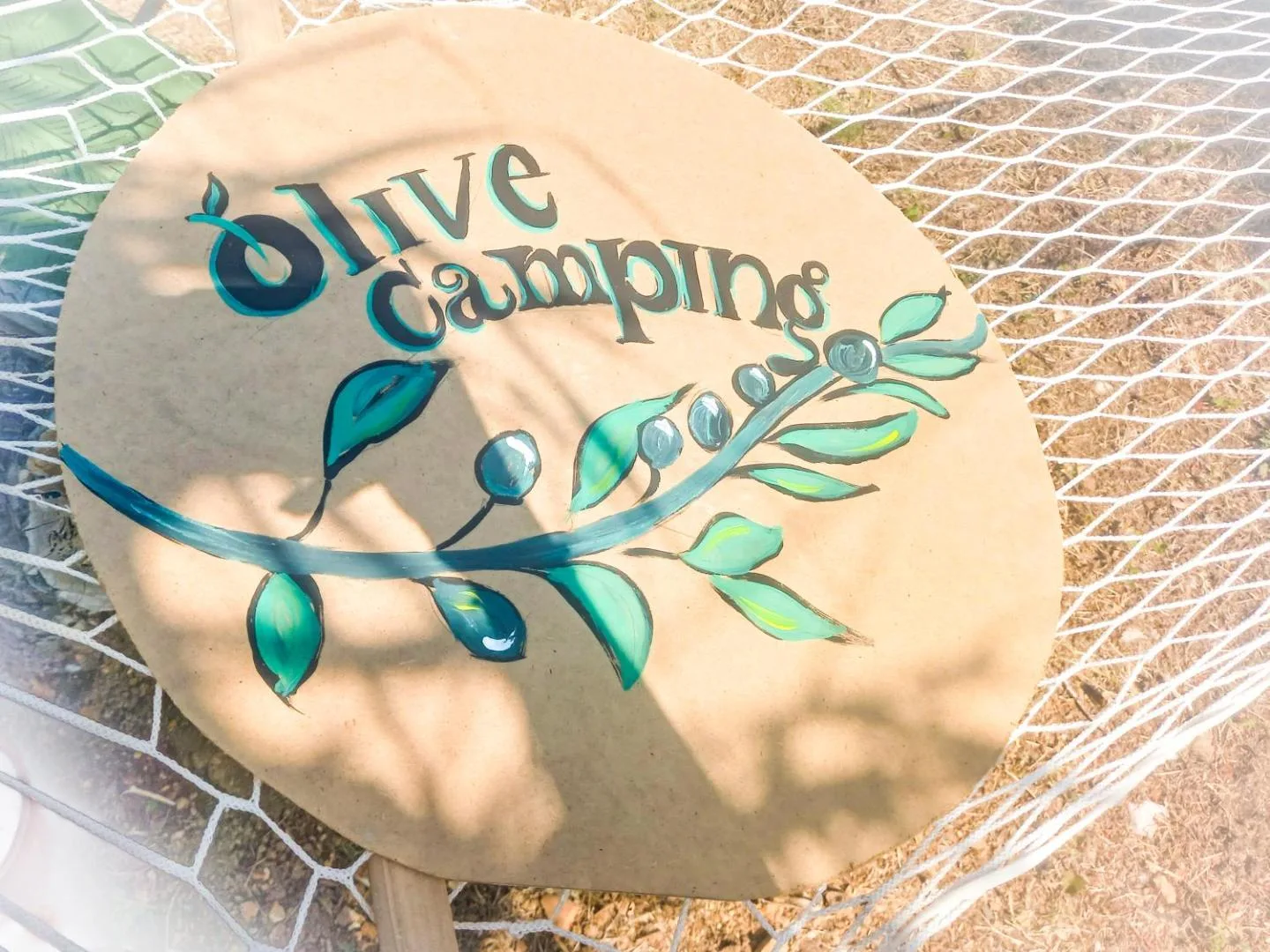 The camp's own hand-painted sign — the words Olive Camping and an olive branch in teal and green on a round board, resting in the netting of a hammock