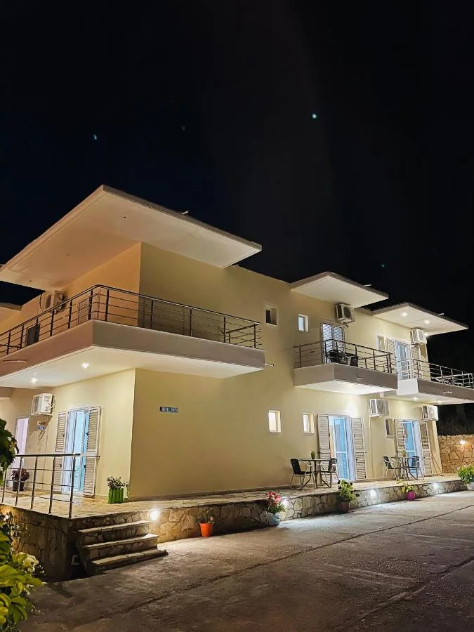 The hotel at night: a rendered two-storey block with deep cantilevered balcony slabs, pale blue shutters and ground-level uplighting