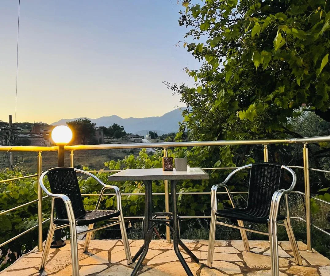Two black bistro chairs and a table on the hotel's flagged terrace at dusk, a globe lamp lit on the balcony rail and the mountains behind